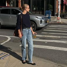 Clara wears a black crewneck sweater, blue jeans, a black belt, black ballet flats, and a brown leather bag.